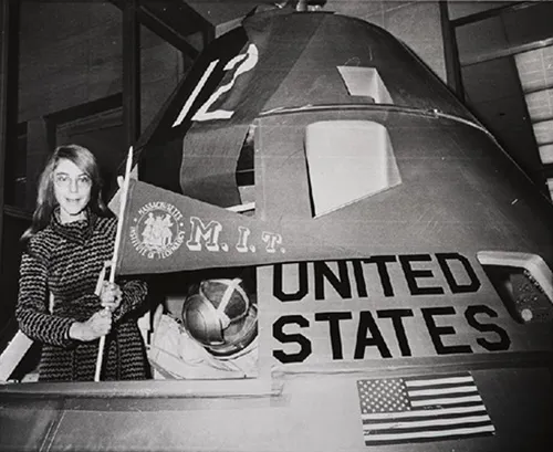 Margaret Hamilton standing in a spacecraft model and holding an M. I. T. flag